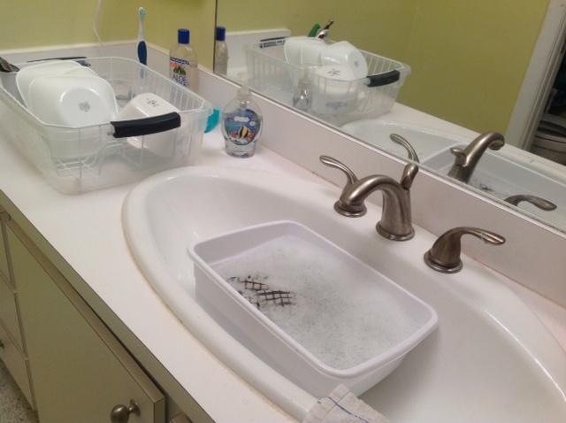 dishpan and dishcloth in a bathroom sink next to dishes in a drainer with clean bowls